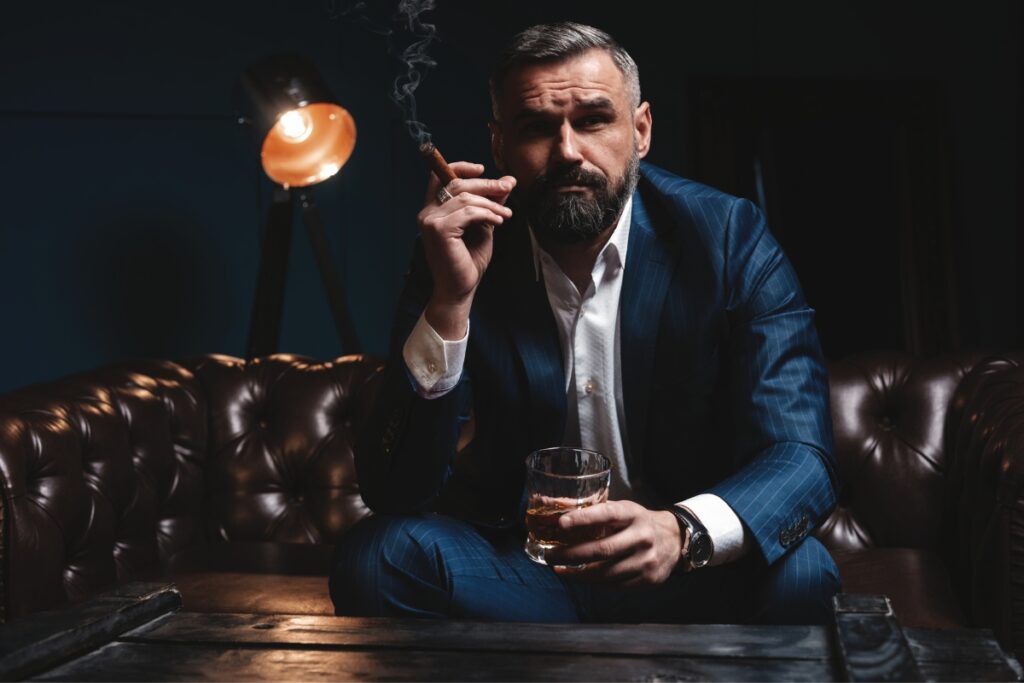 A bearded man sitting on a couch with a glass of whiskey, enjoying a cigar.