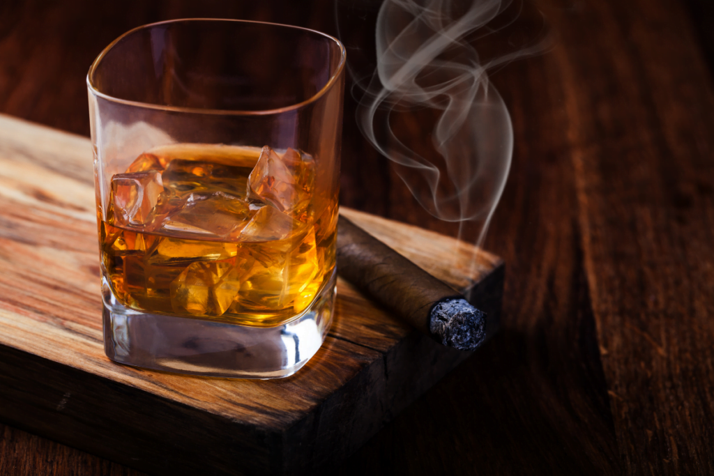 A glass of whiskey with a cigar on a wooden cutting board.