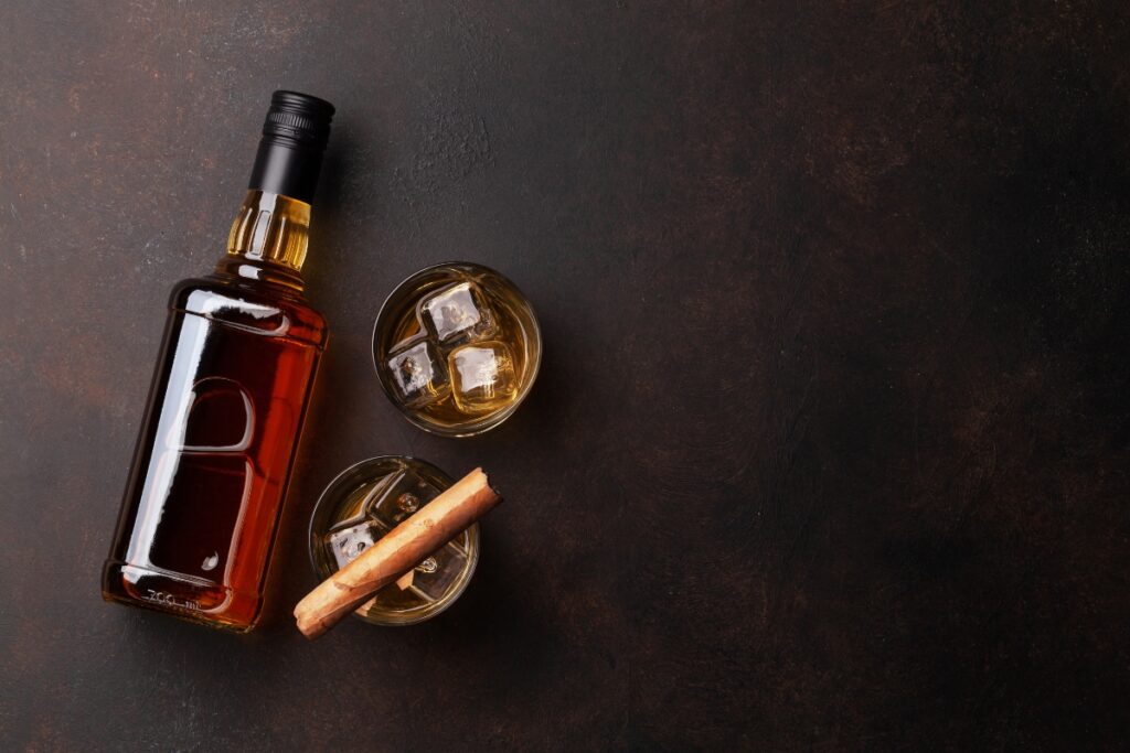 A bottle of whiskey with ice, cinnamon sticks, and a cigar on a dark background perfect for whiskey pairings.
