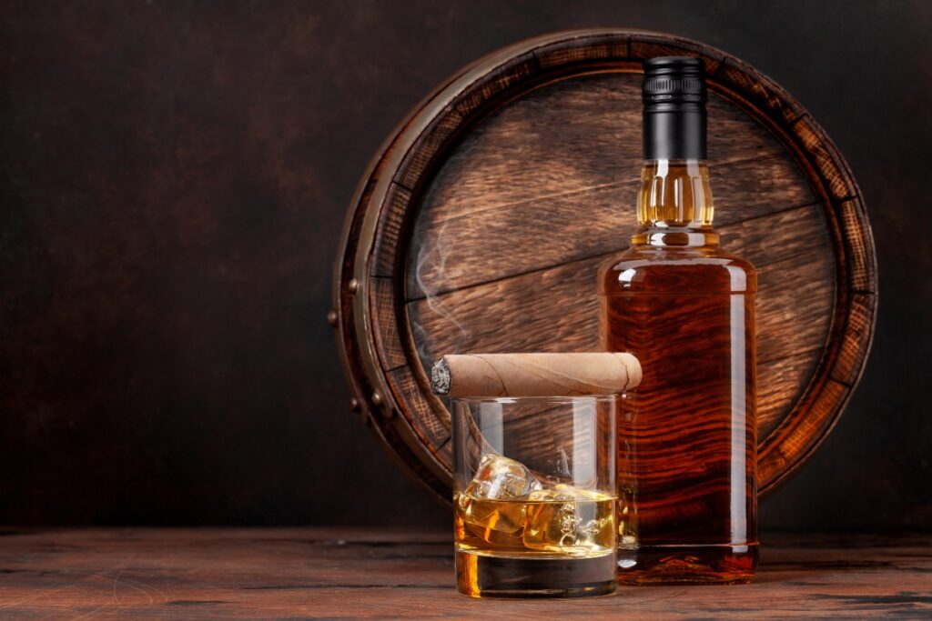 A bottle of whiskey and a glass on a wooden barrel, perfect for cigar and whiskey pairings.