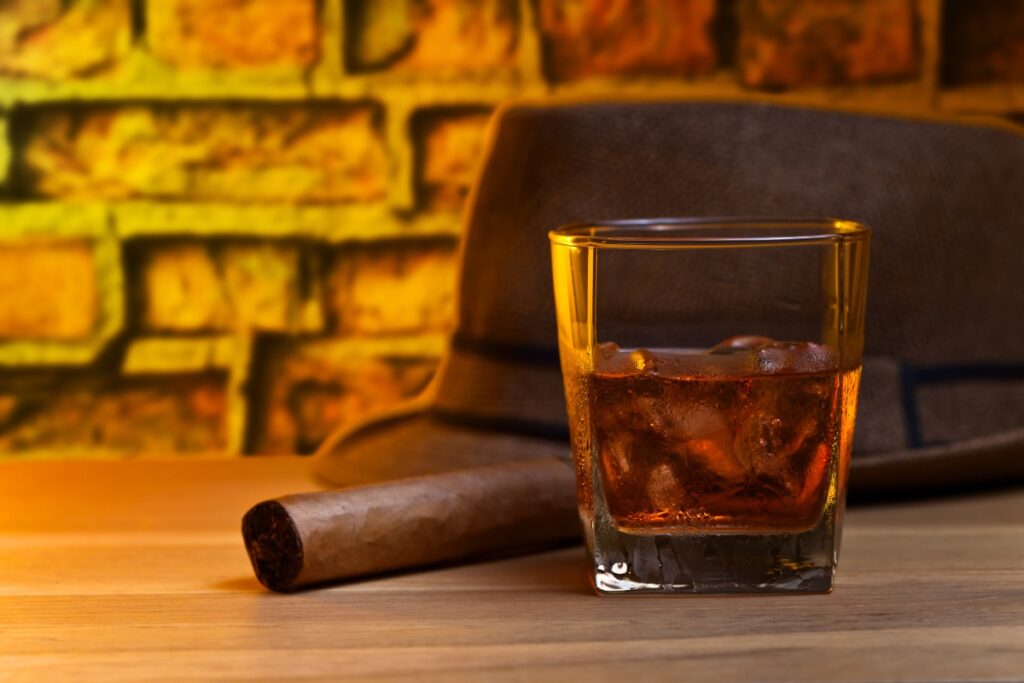 A glass of whiskey and a cigar, perfect for whiskey pairings, rest beside a hat on a wooden table.