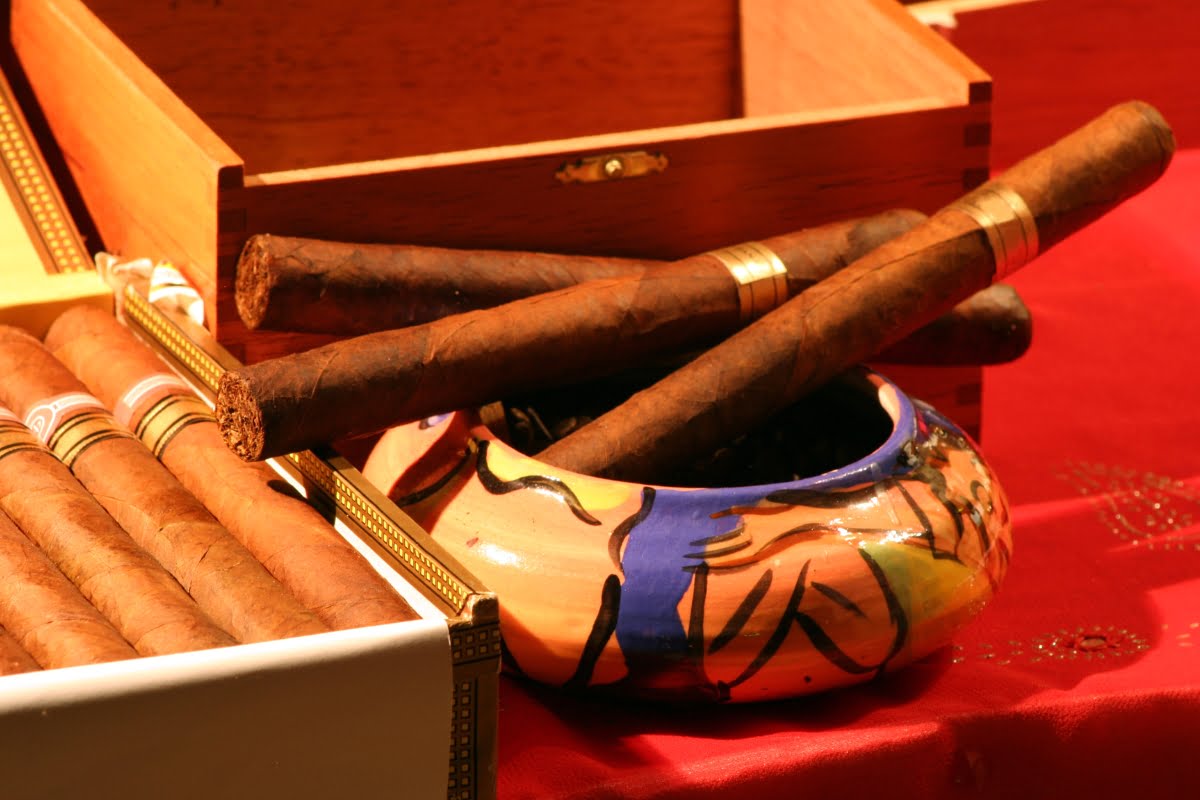 Hand-rolled cigars with distinct flavors displayed in an open wooden box alongside a colorful ceramic ashtray.