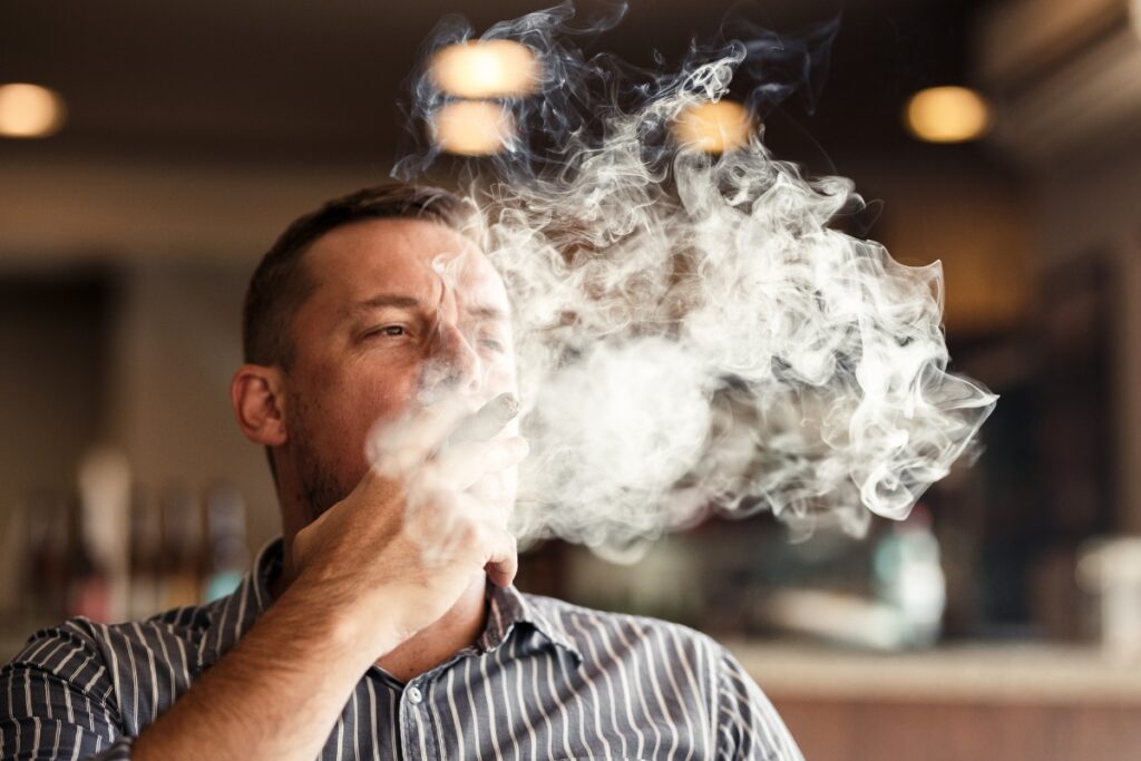 Man practicing cigar etiquette by exhaling smoke indoors.