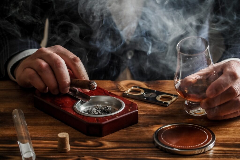 A person practicing cigar etiquette while enjoying a cigar with a glass of whiskey and smoking accessories on a wooden table.