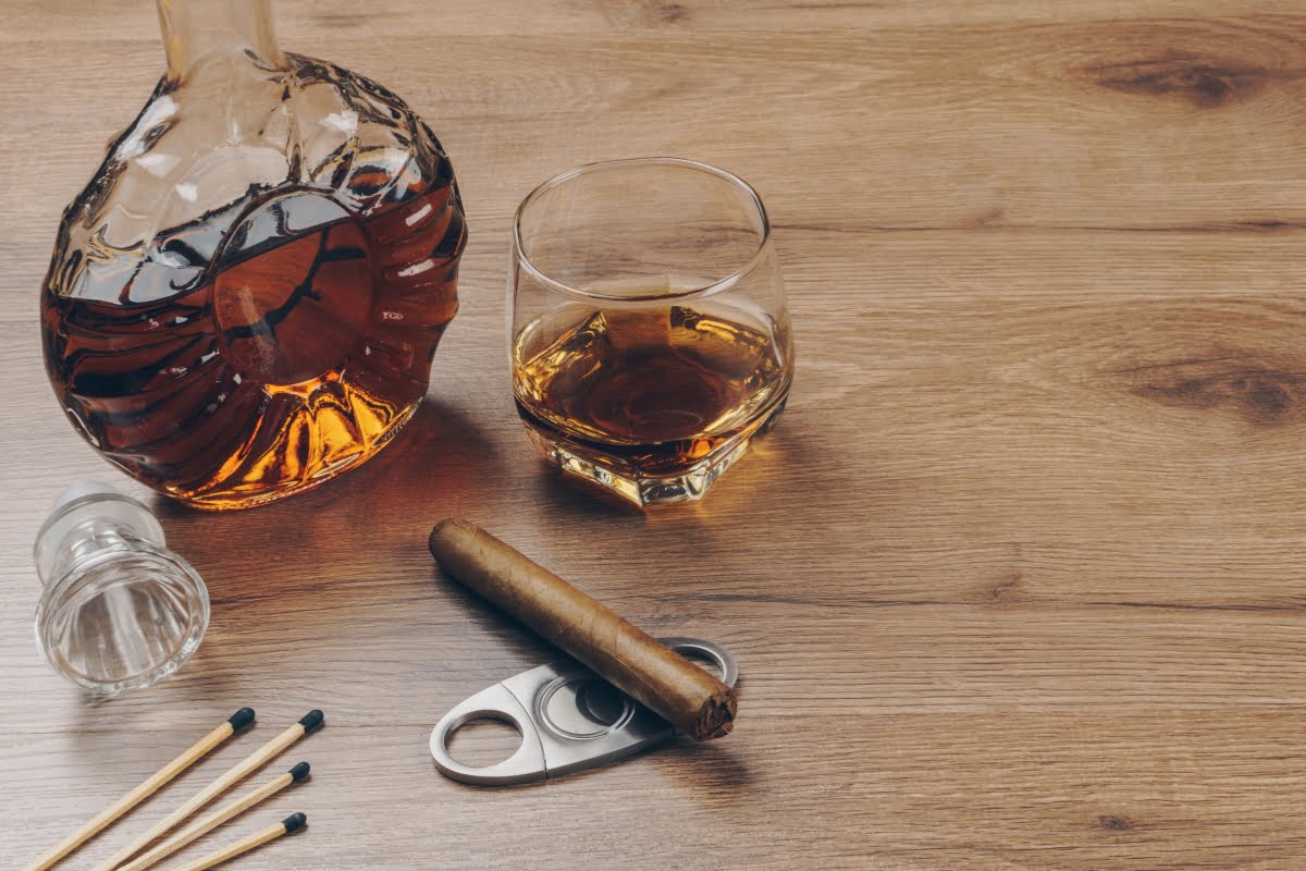 A decanter and a glass of whiskey with the latest cigar trends and a cutter on a wooden table.