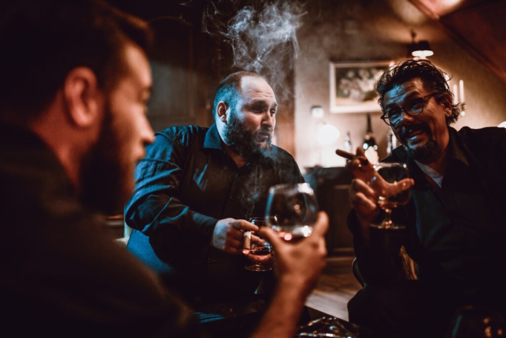 Three men enjoying a conversation over drinks in a dimly lit room, with one exhaling cigar smoke and gesturing animatedly.