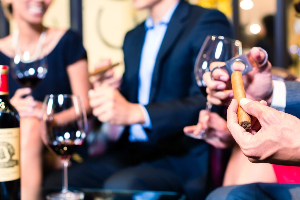 Close-up of a man's hand holding a cigar at cigar events with a blurred background of two people holding wine glasses in a bar.