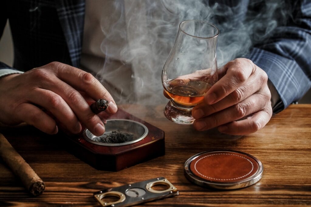 A person holds a cigar and a glass of whiskey, with smoke rising around their hands, sitting at a wooden table with cigar event accessories.