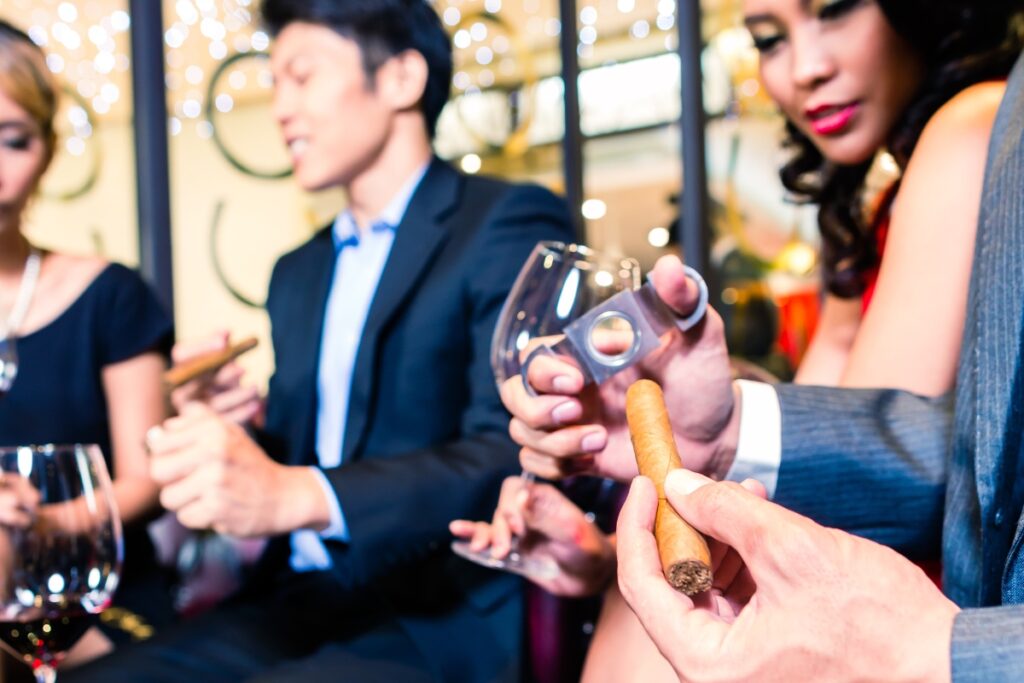 A group of elegantly dressed adults socializing at a cigar event, with a focus on a person holding a cigar and a wine glass.