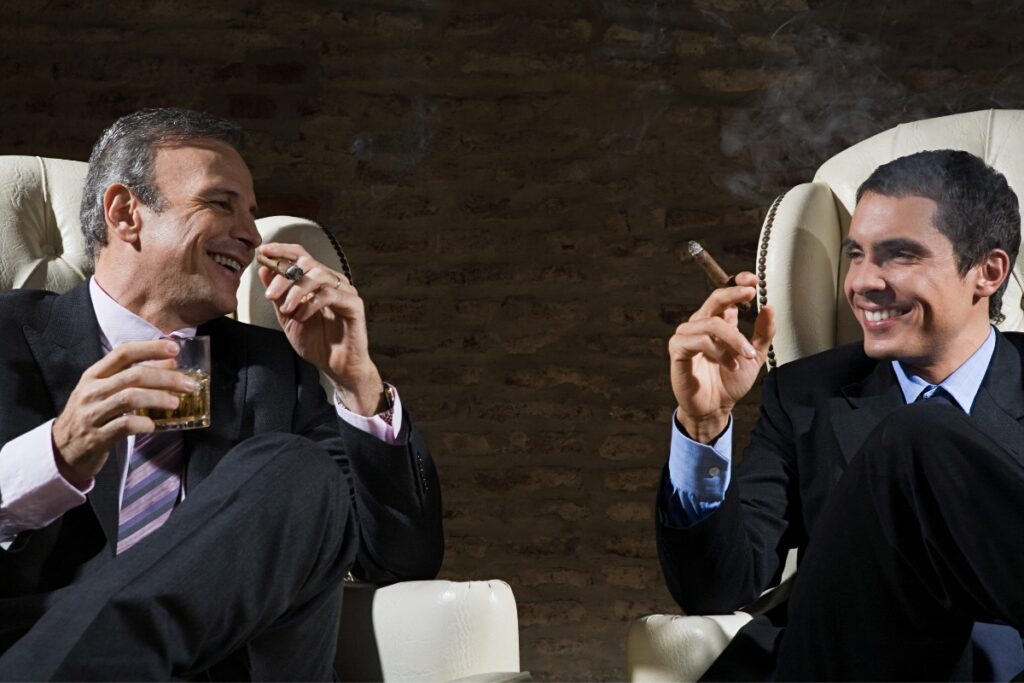 Two men in suits sitting, laughing, and enjoying a cigar event with drinks in hand, in front of a brick wall.