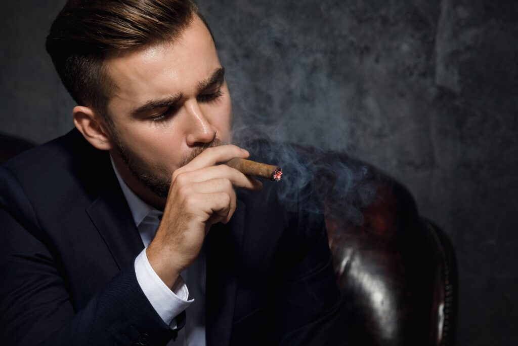 A well-dressed man in a suit sitting on a leather chair, attending a cigar event with smoke swirling around, against a dark gray background.