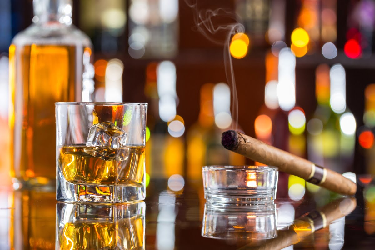 A glass of whiskey with ice on a bar counter alongside a lit cigar, emphasizing health and safety in cigar smoking, with colorful bokeh lights in the background.