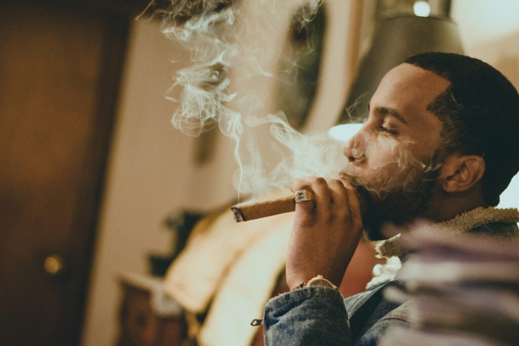 A man smoking a cigar indoors, exhaling smoke, with soft lighting and blurred background, disregarding health and safety in cigar smoking.