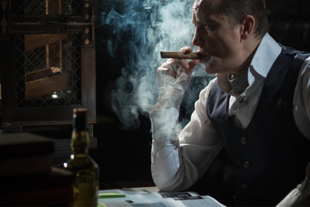 A man in a vest and tie smokes a cigar, examining health and safety in cigar smoking, with a bottle of liquor and a book beside him in a dimly lit room.