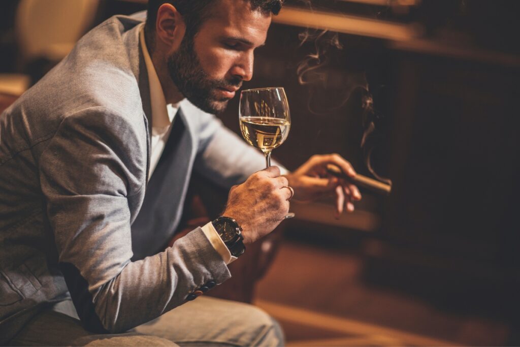 A man in a suit holds a cigar, considering health and safety in cigar smoking, and a glass of white wine, contemplating thoughtfully in a dimly lit setting.