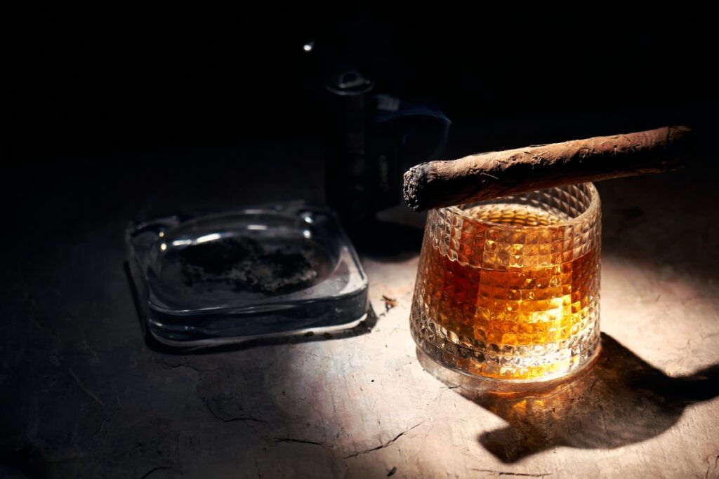 A glass of whiskey with a cigar on top, beside an ashtray and camera on a dark, textured surface, with dramatic lighting highlighting the health and safety concerns in cigar smoking.