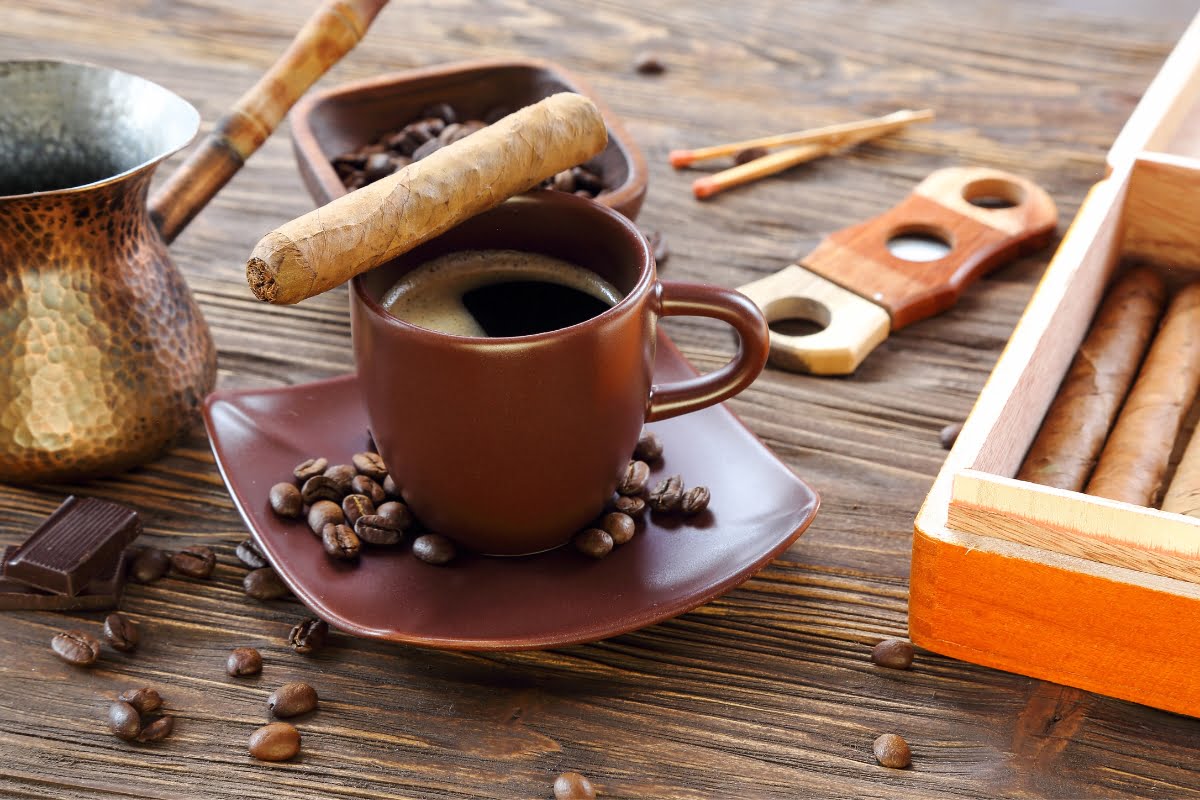 A cup of black coffee sits on a wooden table surrounded by coffee beans, next to a cigar, a cezve pot, and a box of cigars—creating the perfect blend of coffee and cigars.