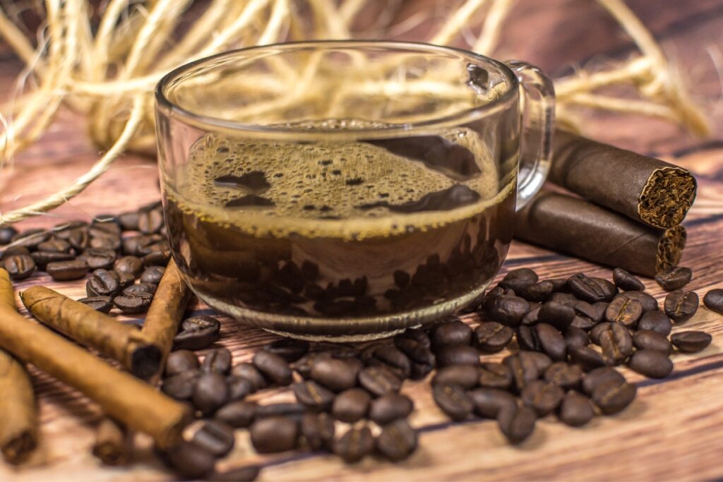 A glass cup of black coffee sits on a table surrounded by coffee beans, cinnamon sticks, and two cigars in the background, creating an enticing scene that perfectly marries coffee and cigars.