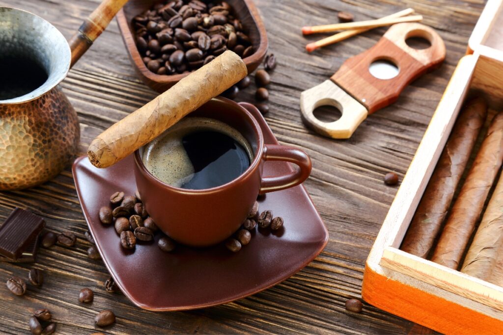 A brown coffee cup with black coffee, a cigar on top, coffee beans scattered around, and a cigar box, cutter, and cezve on a wooden table—a perfect harmony of coffee and cigars.