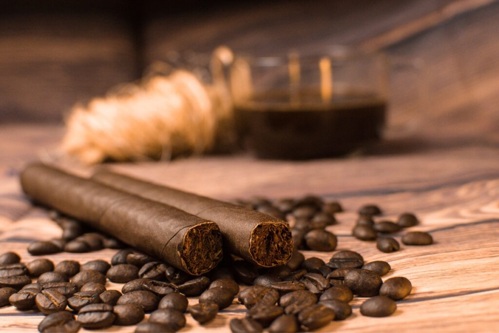 Two cigars rest on a wooden surface surrounded by coffee beans, with a glass of coffee and cigars in the background.