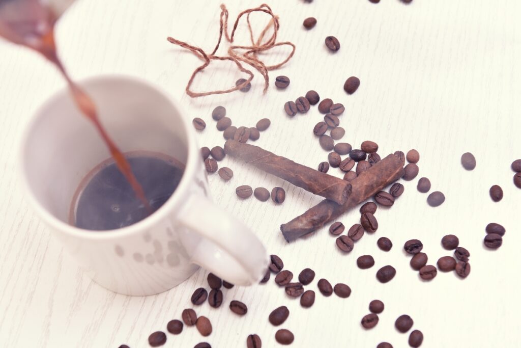 Coffee is being poured into a white ceramic mug on a light surface, surrounded by scattered coffee beans and cinnamon sticks. A piece of twine and a few cigars are visible in the background, adding an air of rustic charm.