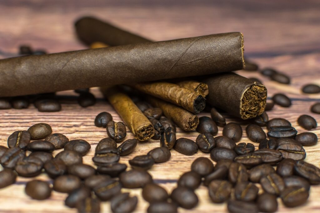 A close-up of coffee and cigars, with beans and rolled leaves scattered across a wooden surface.