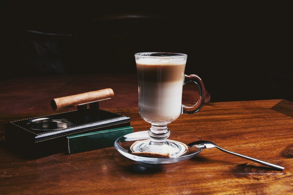 A glass mug of latte on a saucer with a spoon and a cinnamon stick, delicately placed on a wooden table next to a cigar on a cigar holder, reflecting the perfect pairing of coffee and cigars.