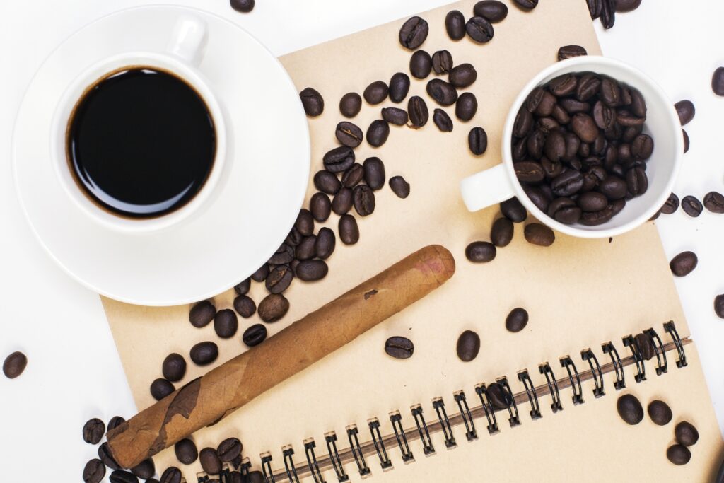 A cup of black coffee, a cup filled with coffee beans, scattered coffee beans, a cigar, and an open notebook are arranged on a light brown surface—a perfect setup for those who appreciate the finer things like coffee and cigars.