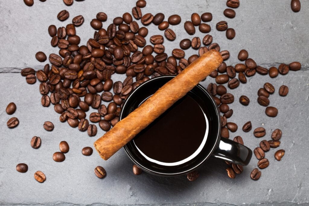 A black coffee mug filled with rich, aromatic coffee, surrounded by scattered beans and complemented by a fine cigar, rests elegantly on a gray surface.