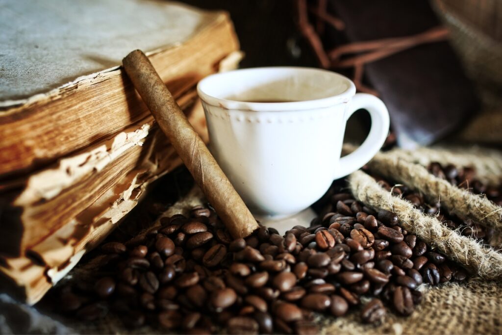 A white coffee cup filled with coffee sits on a surface surrounded by coffee beans, an old book with worn pages, burlap twine, and the comforting aroma of coffee and cigars.