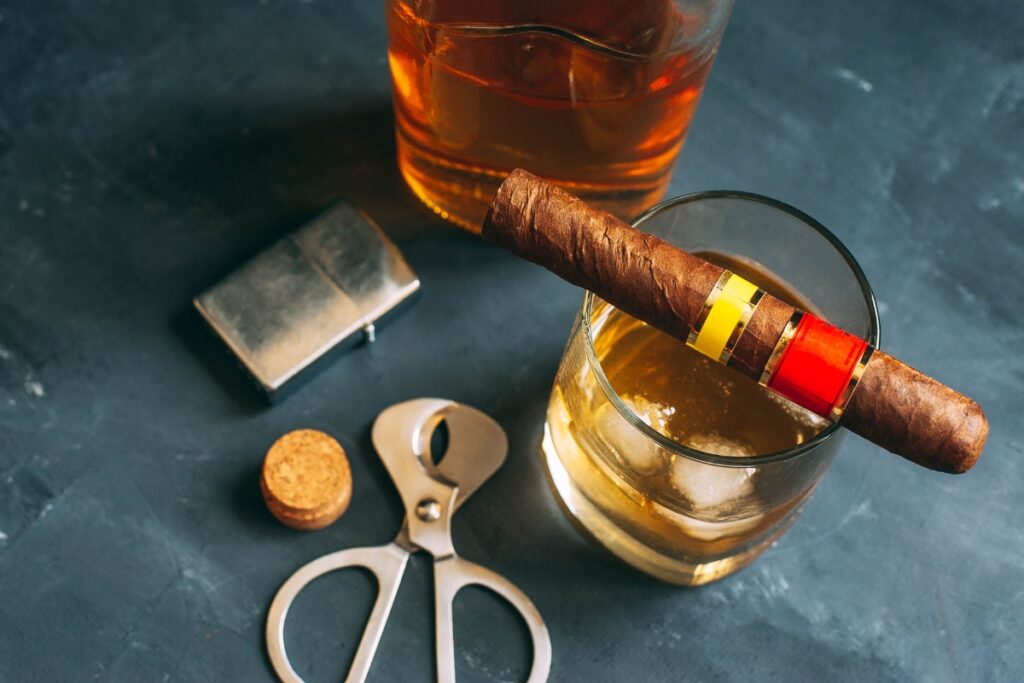 A lit cigar rests on a glass of whiskey. Nearby, luxury cigar accessories like a lighter, cigar cutter, and cork sit elegantly on the dark surface.