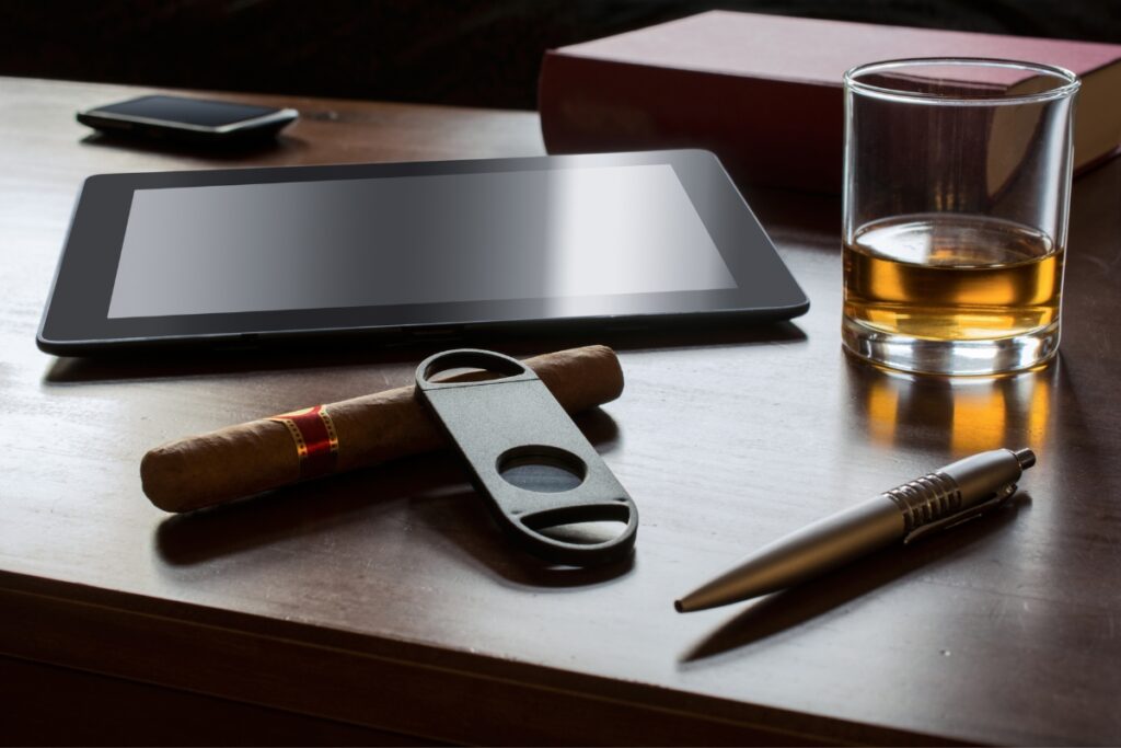 A tablet, an array of luxury cigar accessories including a cigar with cutter, a pen, a glass of whiskey, a book, and a smartphone are elegantly arranged on a wooden surface.