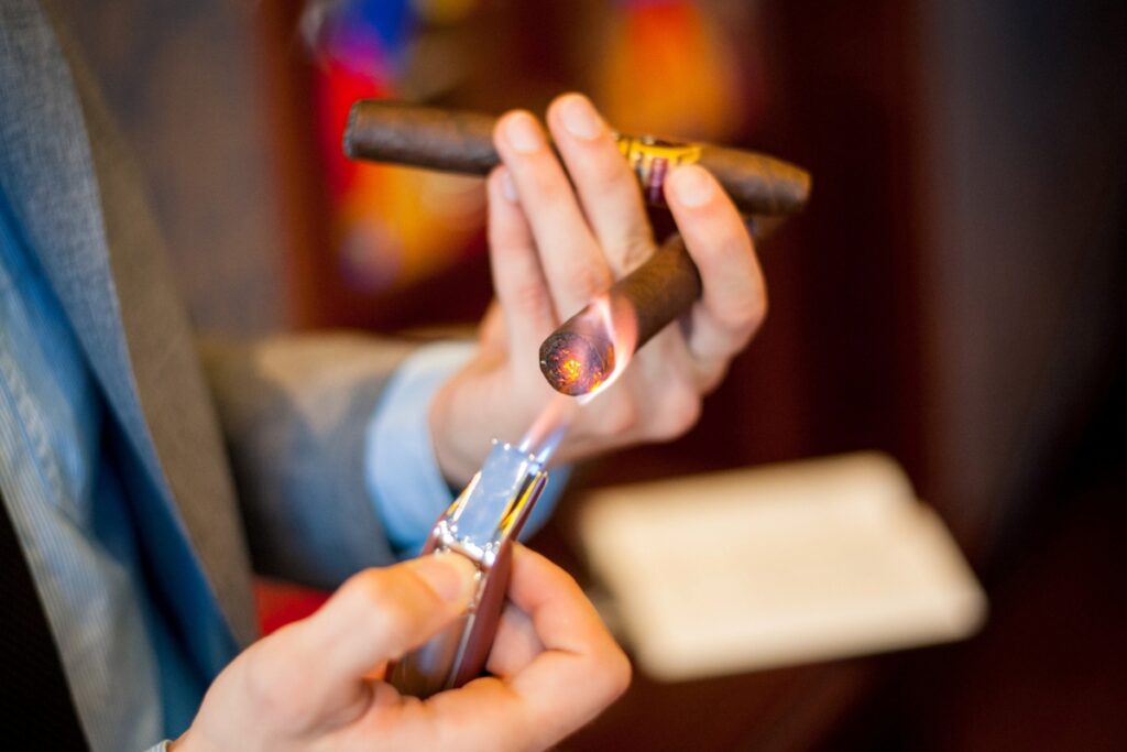 A person in a suit lights a cigar with a metal lighter, showcasing luxury cigar accessories.