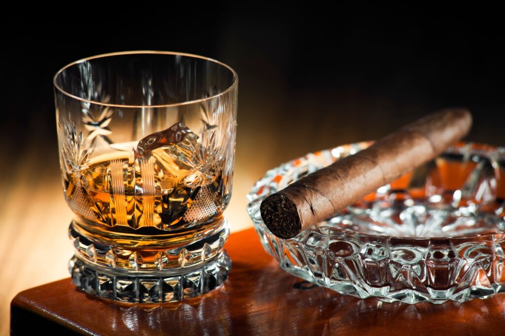 A crystal glass filled with whiskey, ice cubes inside, sits on a wooden surface next to a clear ashtray holding an unlit cigar. The scene is completed by luxury cigar accessories that elevate the experience.