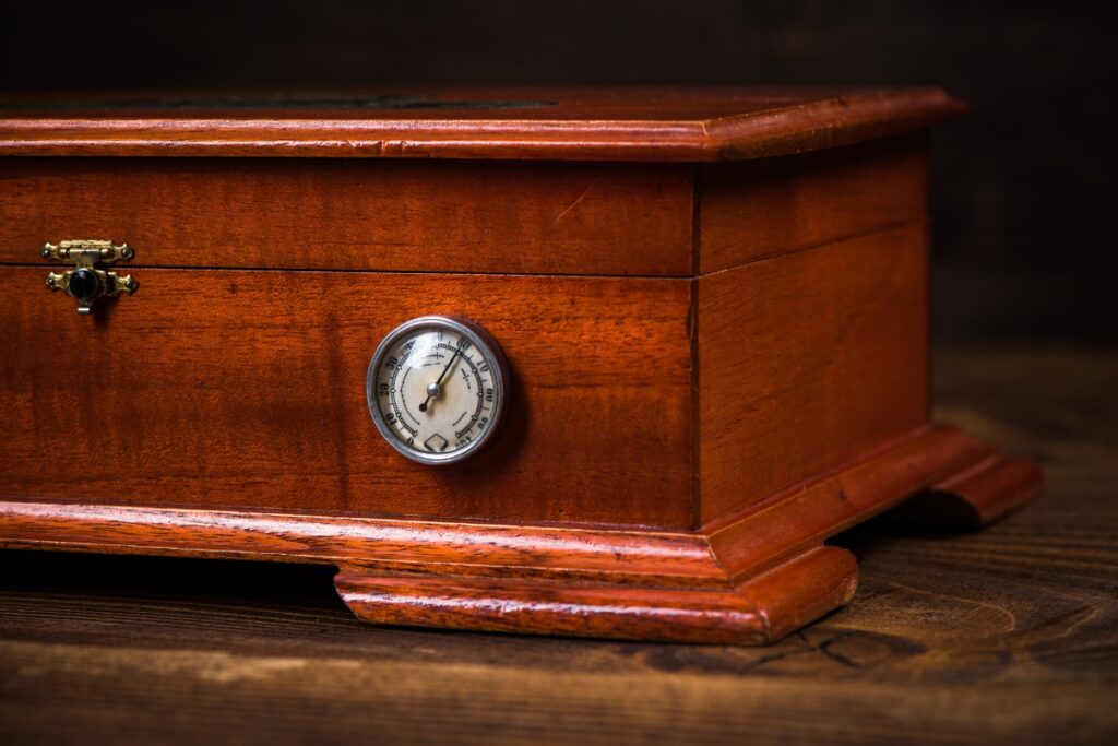 A wooden humidor, a staple of luxury cigar accessories, with a round analog hygrometer on the side sits elegantly on a wooden surface.