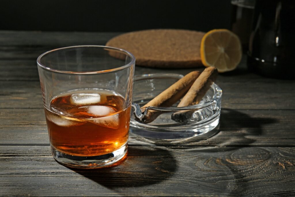 A glass of whiskey with ice sits on a wooden table next to an ashtray holding two luxury cigars, with a slice of lemon in the background.