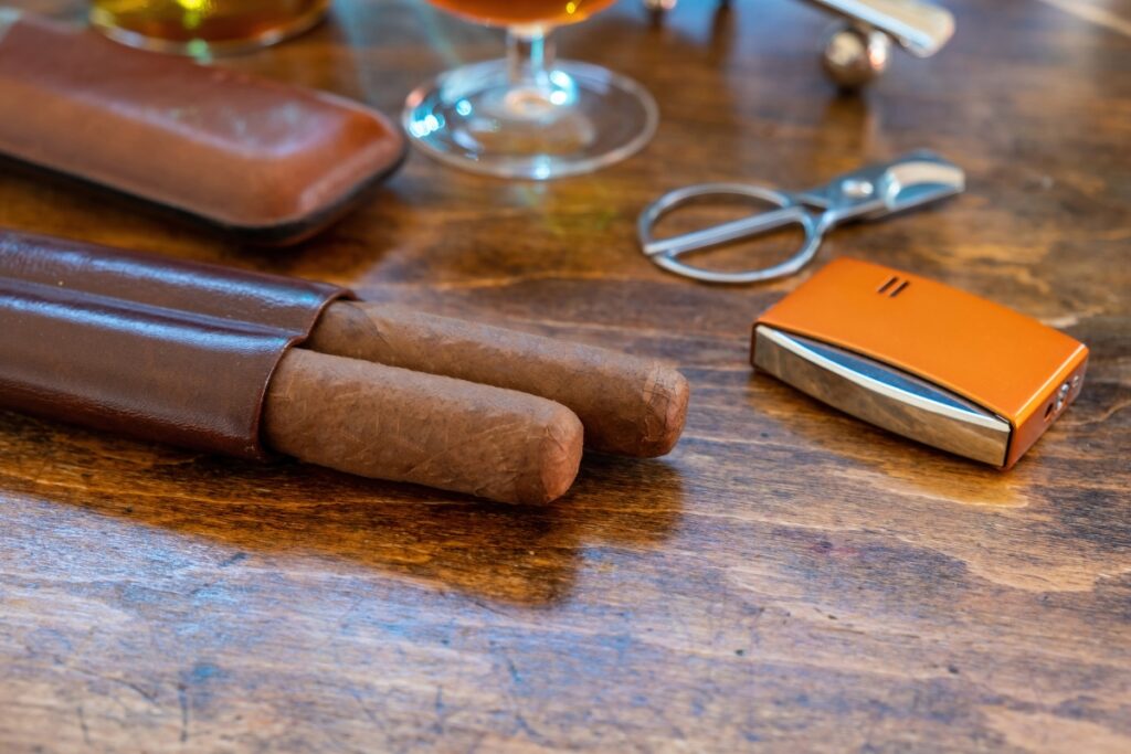 Two cigars in a leather case, a drink in a glass, a pair of cigar scissors, and a lighter placed on a wooden surface – the epitome of luxury cigar accessories.