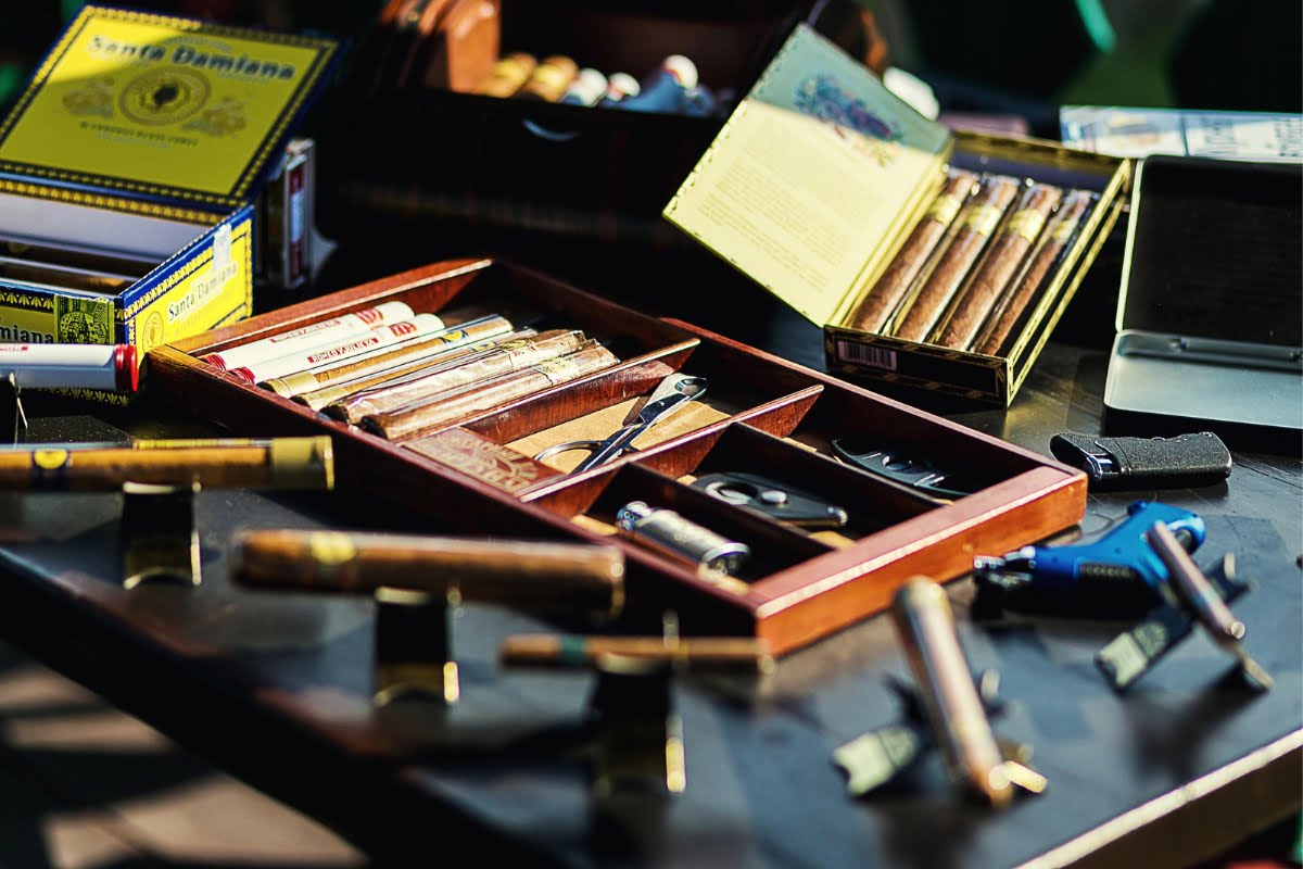 A collection of cigars, cigar cutters, and torches are arranged on a table, perfect for the avid cigar collecting enthusiast. There are several boxes and cases of cigars in the background.