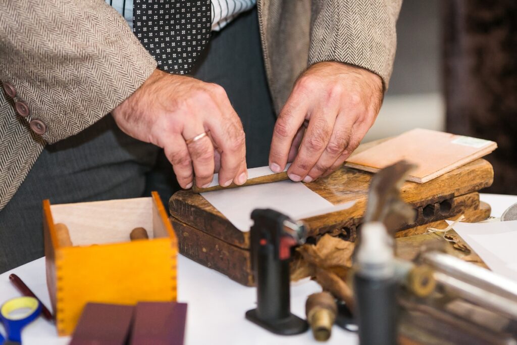 A person rolls a cigar at a table, holding a cigar wrapper around the filler. Various tools and materials, essential for cigar collecting enthusiasts, including parchment papers and a small blowtorch, are placed on the table.