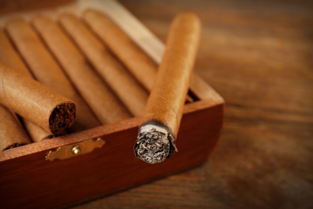 A wooden box containing multiple cigars, with one partially smoked cigar resting on the edge of the box against a wooden background, showcases the art of cigar collecting.