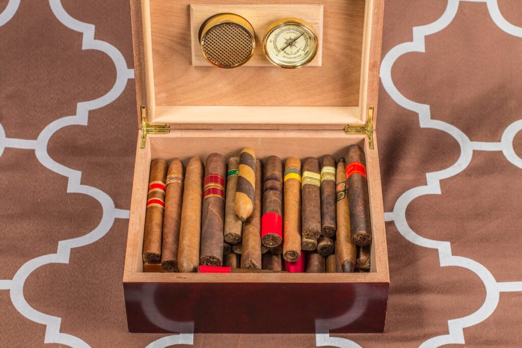 A wooden humidor box, perfect for cigar collecting, contains an assortment of cigars of various sizes and bands. It features a built-in hygrometer gauge on the lid's interior.