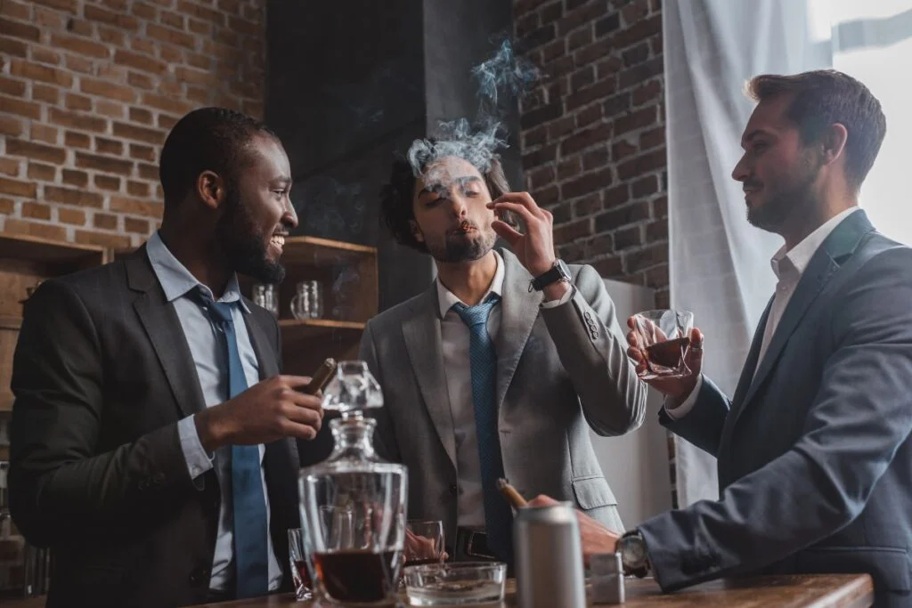 Three men in suits are standing, laughing, and smoking cigars while holding glasses of whiskey in a room with exposed brick walls. A decanter is on the table in front of them as they discuss their passion for cigar collecting.