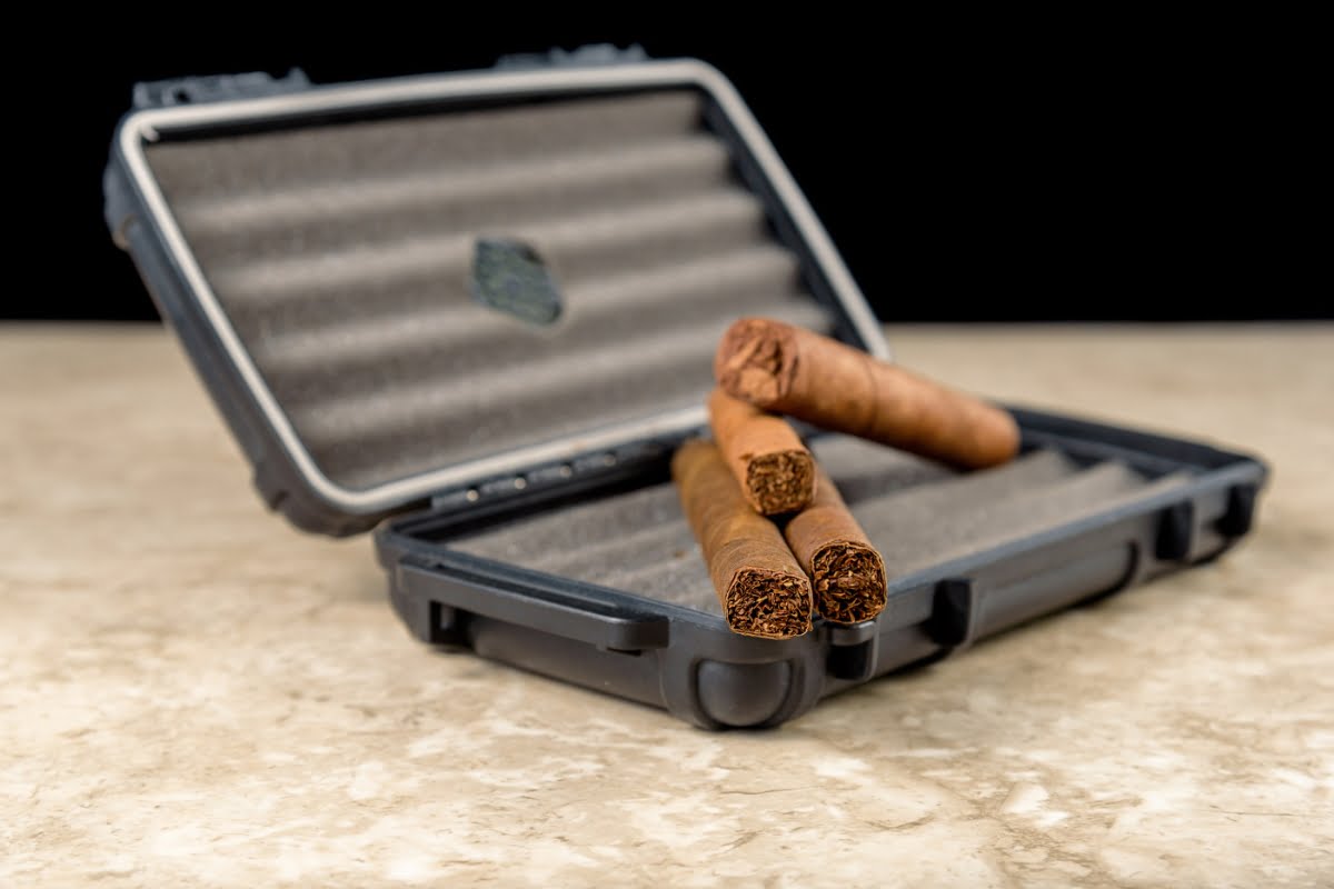 A black travel humidor with foam padding is open, displaying three brown cigars resting on the foam lining on a marble surface.