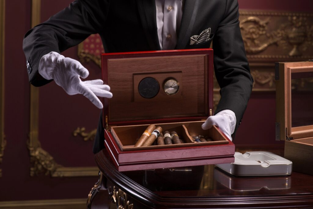 A person in white gloves and formal attire opens a wooden cigar humidor on a polished wooden table, revealing several cigars inside, alongside compact travel humidors for connoisseurs on the go.