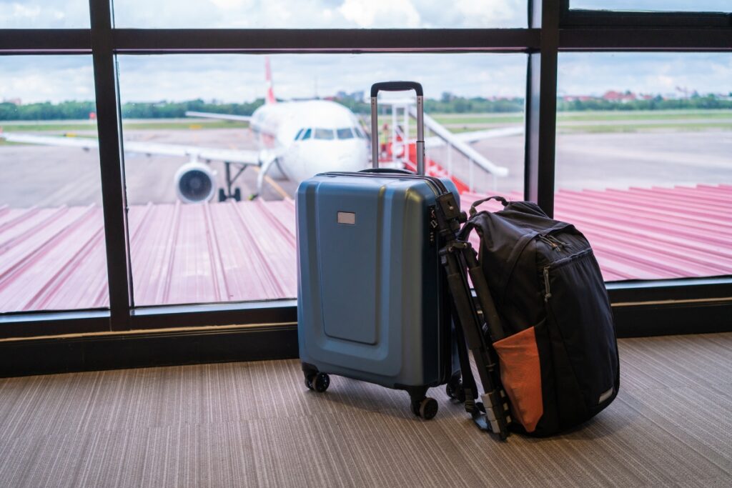 A blue suitcase and a black backpack, with travel humidors tucked inside, are placed on the floor near a large window overlooking an airplane parked at the gate.