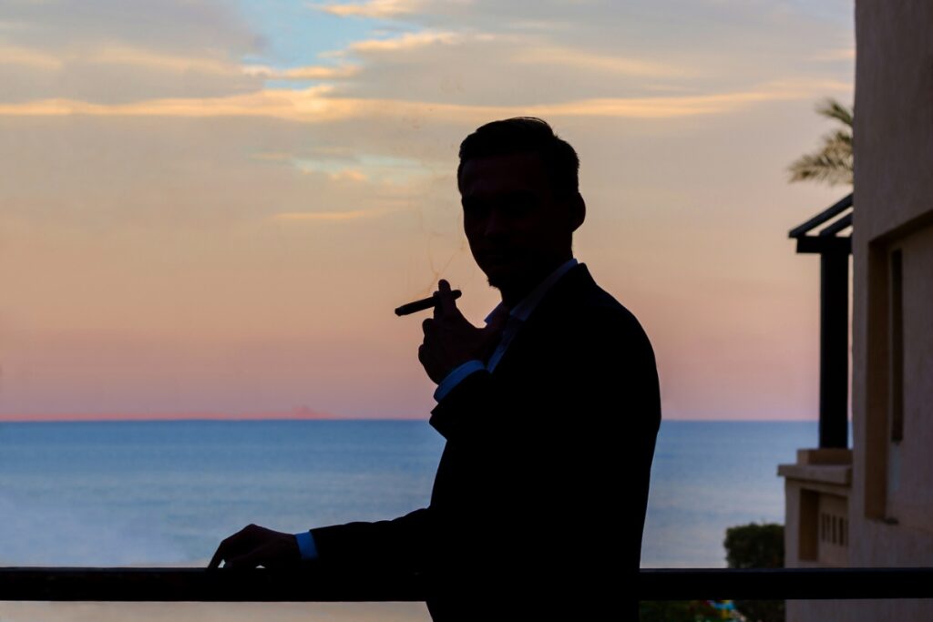 Silhouette of a person in a suit holding a cigar and standing on a balcony with a view of the ocean and a colorful sunset sky, showcasing an elegant travel humidor by their side.