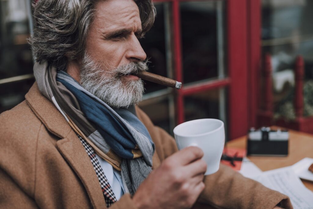 A man with gray hair and a beard, dressed in a brown coat and scarves, holding a white cup in one hand and a cigar from his travel humidor in his mouth, sits at an outdoor cafe.