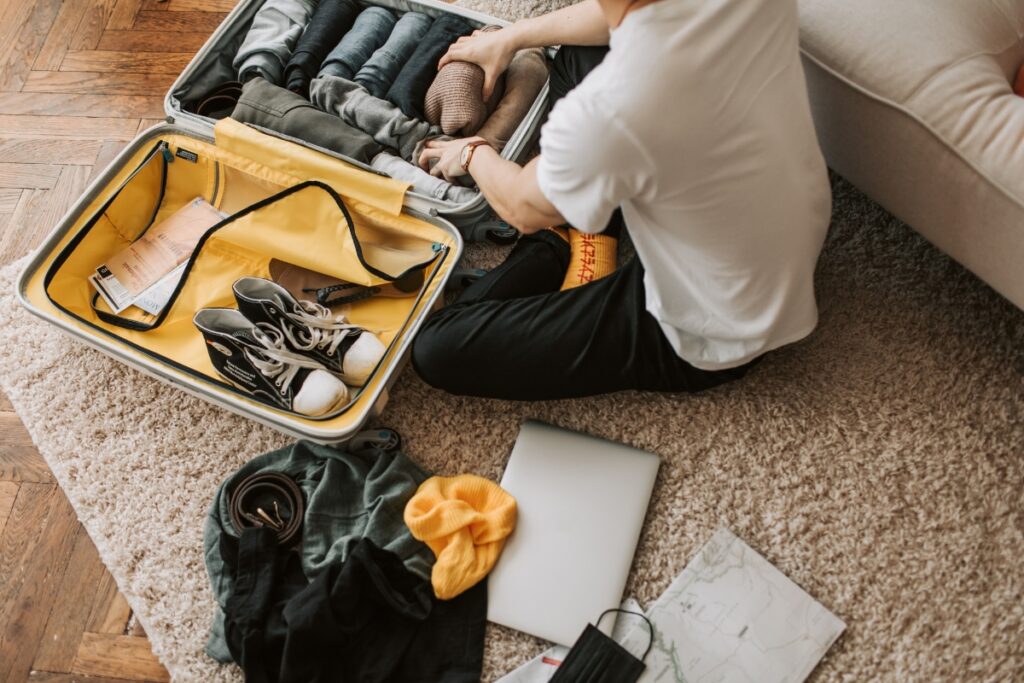 Person sitting on a beige carpet, packing a suitcase with rolled clothes, shoes, travel items—including a laptop, hat, map, and travel humidors.