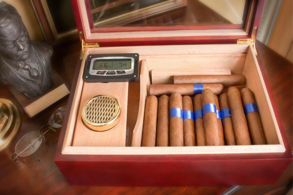 A wooden humidor containing cigars with blue bands, a hygrometer showing humidity and temperature, a metal ventilation cover, a pair of glasses, and a sculpture in the background evoke an elegant scene. Perfect for those who might need travel humidors on their journeys to maintain their collection.