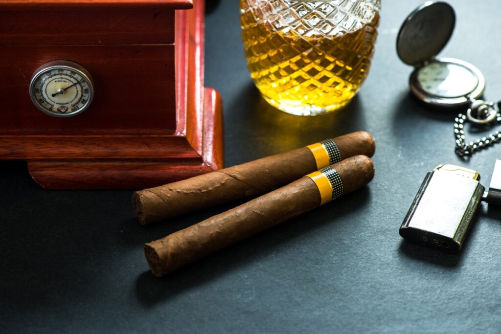 Two cigars, a glass container of amber liquid, a pocket watch, a lighter, and a wooden box with a small clock are arranged on the dark surface. Nearby, sleek travel humidors ensure your cigars remain in perfect condition while on the go.